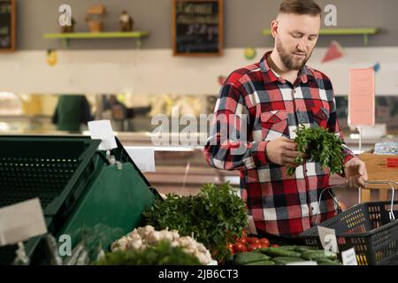 Junger kaukasischer Mann, der Gemüse und Grün im Supermarkt, in der Gemüseabteilung und beim Einkaufen von Männern kauft Stockfoto