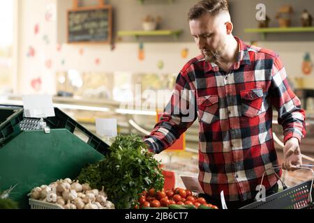 Junger kaukasischer Mann, der Gemüse im Lebensmittelbereich im Supermarkt kauft. Wählen Sie Gemüse und Gemüse, während Sie den Lebensmittelkorb halten. Tausendjähriger Mann Stockfoto