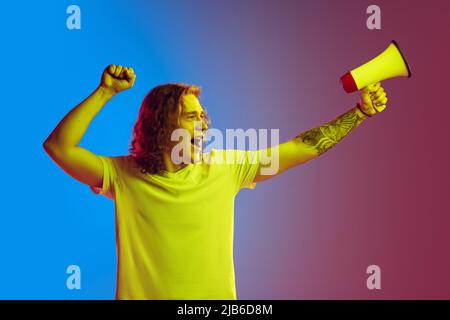 Junger aufgeregt Mann mit langen lockigen Haaren schrie auf Megaphon isoliert auf gradienten blau-rosa Hintergrund. Konzept der Schönheit, Mode, Jugendkultur und Stockfoto