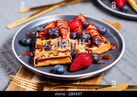 Belgische Waffel mit Schokolade, Erdbeere, Heidelbeeren und Puderzucker auf dunklem Teller Stockfoto