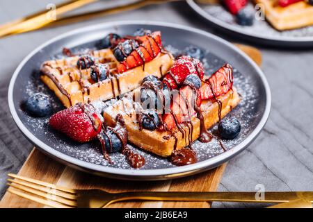 Belgische Waffel mit Schokolade, Erdbeere, Heidelbeeren und Puderzucker auf dunklem Teller Stockfoto