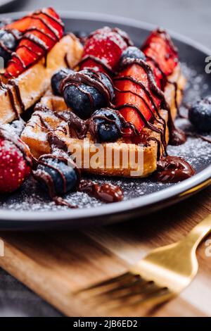 Belgische Waffel mit Schokolade, Erdbeere, Heidelbeeren und Puderzucker auf dunklem Teller Stockfoto