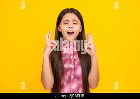 Froh Teenager-Mädchen kreuzt die Finger, schließt die Augen mit Vergnügen, erwarten hören gute Nachrichten, isolierten gelben Hintergrund. Glückliches Kind freut sich, dass seine Stockfoto