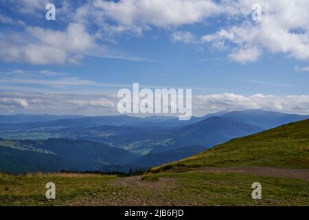 Breiter Feldweg, der vom Hügel mit trockener Vegetation im Frühherbst absteigt. Blick über den Wanderweg, der den Hang hinunter führt, mit Panoramablick auf den Bergrücken im Hintergrund. Konzept des Reisens. Stockfoto