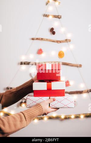 Die Hände der Frau halten Geschenkschachteln gegen die Dekoration Weihnachtsbaum aus trockenen Sticks, trocken orange, Zapfen auf weißem Hintergrund. Zero Waste Christmas, Stockfoto