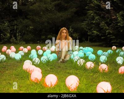 Eine Frau sitzt auf grünem Rasen zwischen vielen leuchtenden bunten Bällen in einem Sommerpark Stockfoto