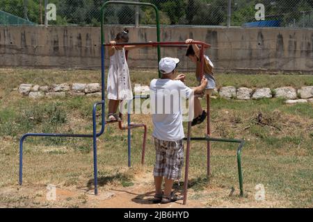 Enkelkinder, die sich am sonnigen Tag auf dem Spielplatz mit dem Großvater amüsieren. Rückansicht älterer Mann, der ihren kleinen Enkel unterstützt, während er an einem schönen sonnigen Tag auf dem Spielplatz im Freien spielt. Stockfoto