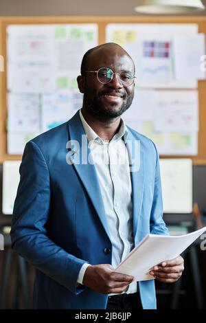 Porträt eines erfolgreichen afrikanischen Geschäftsmannes in blauem Anzug, der Dokumente hält und lächelt, wenn die Kamera im Büro steht Stockfoto