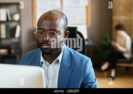 Seriöser afrikanischer Geschäftsmann in Brillen und blauem Anzug, der im Büro am Computer arbeitet Stockfoto