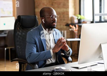 Afrikanischer selbstbewusster Geschäftsmann in Anzug, der auf einem Stuhl an seinem Arbeitsplatz vor einem Computermonitor sitzt und mit einem Kollegen online spricht Stockfoto