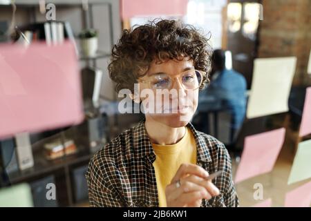 Junge Geschäftsfrau mit lockigen Haaren in einer Brille, die Notizen auf Aufklebern macht, die an der Glaswand im Büro hängen Stockfoto