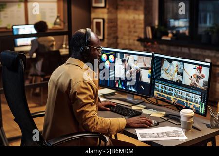 Afrikanischer junger Kolorist, der Fotos mit Software am Computer bearbeitet, während er am Tisch sitzt und bis spät in die Nacht im Büro arbeitet Stockfoto