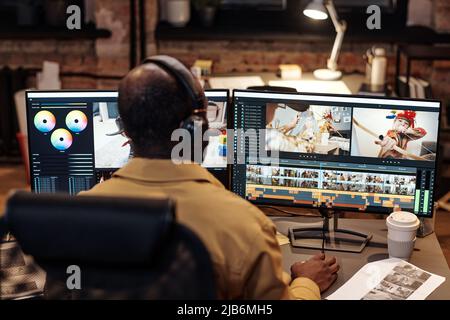 Rückansicht des Redakteurs oder Videodesigners in Kopfhörern, die vor dem Computerbildschirm sitzen und mit neuen Inhalten arbeiten Stockfoto