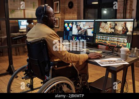 Rückansicht des Redakteurs in Kopfhörern, der im Rollstuhl vor dem Computerbildschirm sitzt und mit neuen Inhalten arbeitet Stockfoto