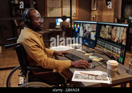 Afrikanischer professioneller Redakteur mit Behinderung, der bis spät in die Nacht am Schreibtisch mit neuen Inhalten am Computer arbeitet Stockfoto