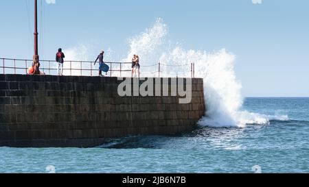 Ein Paar wird von einer Welle erwischt, die über dem Porthleven Pier in Cornwall zusammenbricht Stockfoto