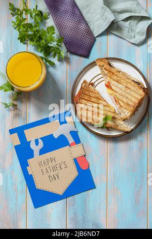 Wir feiern den Vatertag. Frühstück. Vatertagskarte und Heimwerker-Sandwich mit Speck auf Holztisch. Blick von oben. Stockfoto