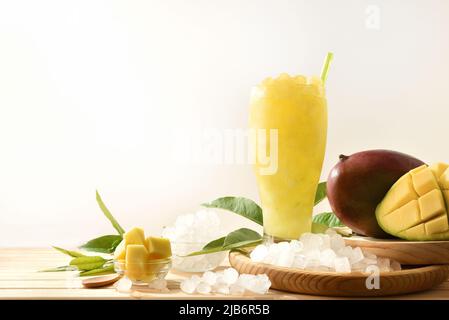 Detail eines Glases mit Mangosaft mit Eis auf einem Holztisch mit Früchten und zerdrücktem Eis um ihn herum. Vorderansicht. Horizontale Zusammensetzung. Stockfoto