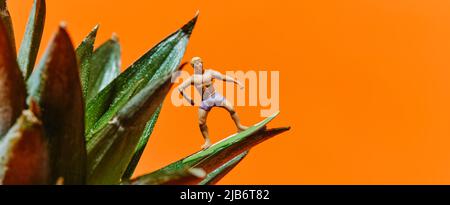 Ein Miniatur-Surfer-Mann, der auf dem Surfbrett auf den Blättern einer Ananas auf einem orangefarbenen Hintergrund mit etwas Leerzeichen rechts in einem Panor reitet Stockfoto