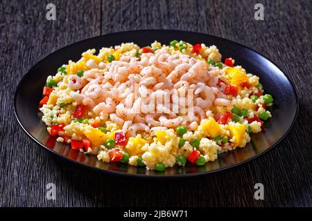 Gekochte kleine rosafarbene Garnelen auf Hirse-Pilaf mit Gemüse, Mangostückchen und Gewürzen auf schwarzem Teller auf dunklem Tisch, horizontale Ansicht von oben Stockfoto