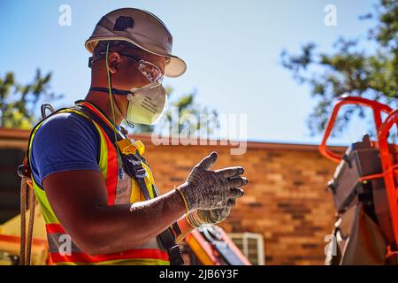 Ein Bauarbeiter, der während des COVID vollständige PSA und Gesichtsmaske trägt, bereitet sich auf den Betrieb eines Hubwerks mit Hubarbeitsbühnen vor. Stockfoto