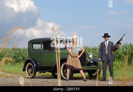 Bonnie und Clyde stehen zu ihrem Fluchtauto. Sie sind mit Waffen bewaffnet und in Kleidung im Stil von 1930 gekleidet. Stockfoto