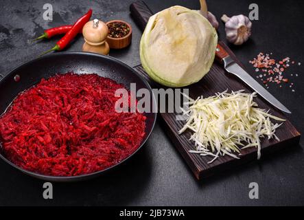 Gehackte Rote Bete in einer Pfanne sowie Gewürze und Kräuter auf einem Holzschneidebrett als Zutaten für die Zubereitung des traditionellen ukrainischen Borschts Stockfoto