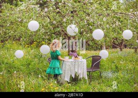 Nettes blondes lockiges Haar Mädchen, das im heimischen Apfelgarten am Tisch mit Teetassen und vielen Papierlaternen steht, die im Frühling vom blühenden Apfelbaum hängen. Stockfoto