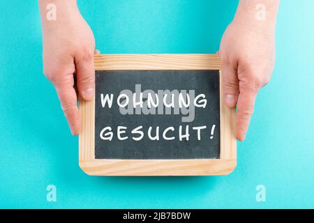 Mit einer Tafel in den Händen steht die Suche nach einer Wohnung in deutscher Sprache auf dem Schild und zieht in ein neues Zuhause, eine Immobilie Stockfoto