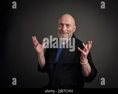 Porträt eines glücklichen, lächelnden und aufgeregten älteren Mannes mit expressiver Handbewegung vor grauem Hintergrund Stockfoto