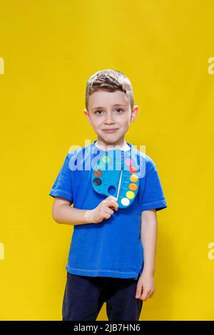 Kleiner netter Vorschuljunge, der eine Palette von Farben und einen Pinsel auf gelbem Hintergrund intensiv hält. Kreatives Kind. Stockfoto