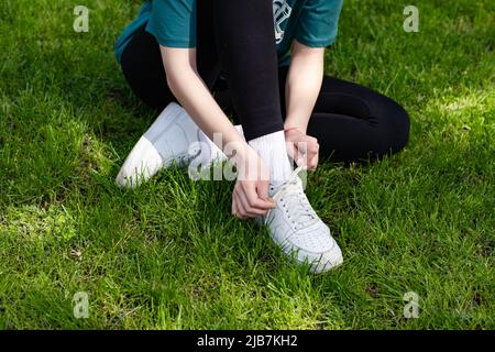 Nahaufnahme einer jungen Rotschopf-Frau mit grünem T-Shirt, die im Stadtpark steht und im Freien Spitzen-Laufschuhe bindet, die sich zum Laufen vorbereiten. Jogging Girl exe Stockfoto