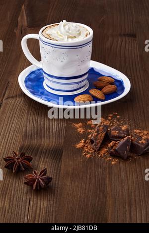 Eine Tasse Cappuccino, gehackte Schokolade, Gewürze und Mandeln auf einem braunen Holztisch. Kaffee-Tag Stockfoto
