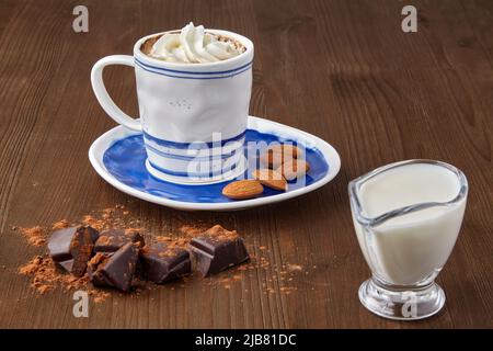 Tasse Kaffee mit Schlagsahne und Mandeln, gehackter Schokolade und Milch in einem Glaskrug Milch auf einem braunen Holztisch. Getränke auf Kaffeebaske Stockfoto