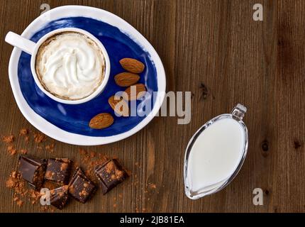 Blaue Tasse Kaffee mit Eis, Mandeln, Schokolade und Milch in einem Milchkännchen aus Glas auf einem dunklen Holztisch Stockfoto
