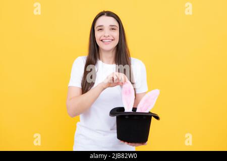 Hasenohren erscheinen aus dem Zauberhut. Porträt eines jungen Mädchens mit Hasenohren isoliert auf gelbem Hintergrund. Stockfoto