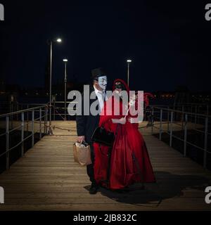 Venedig, Italien - 18.02.2022: Ein Mann und eine Frau in venezianischen Karnevalskostümen stehen nachts auf dem hölzernen Pier am Canal Grande in Venedig Stockfoto