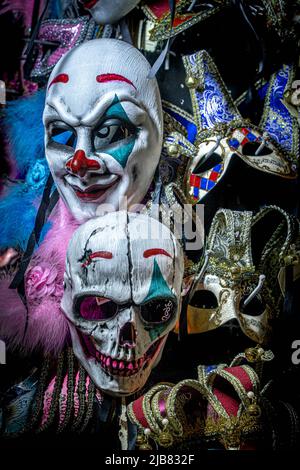 Venedig, Italien - 18.02.2022: Nahaufnahme der furchteinflößenden Clownsmaske und der Totenkopfmaske, die zwischen traditionellen venezianischen Masken zusammenhängen. Frohe Halloween. Stockfoto