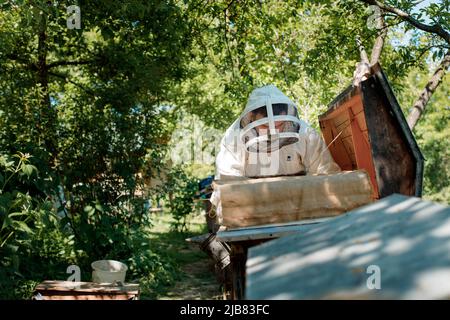 Imker im Schutzanzug arbeitet mit Waben. Ein Bauer im Bienenanzug arbeitet mit Waben in einem Bienenhaus. Imkerei auf dem Land. Ökologischer Landbau. Stockfoto