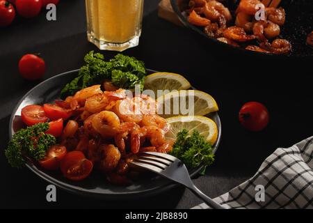 Abendessen serviert: Gebratene Garnelen mit Zitrone, Tomaten und Kräutern sowie Bier in einem Glas auf dem Tisch Stockfoto