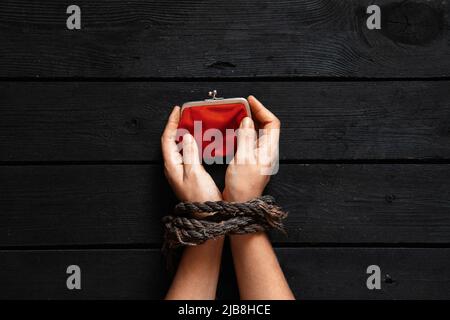 Ein Mädchen mit gefesselten Händen hält eine rote Brieftasche auf einem schwarzen Holztisch, eine Brieftasche in ihren gefesselten Händen Stockfoto