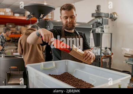 Der Barista verpackt geröstete Kaffeebohnen in Verpackungen, die vor dem Hintergrund der Röstermaschine zum Verkauf stehen Stockfoto