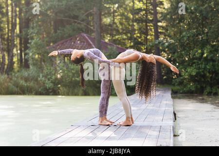 Zwei weibliche Yoga-Praktizierende halten sich gegenseitig bei der Hasta Uttanasana in einer Hinterbeine-Haltung und stehen auf einer hölzernen Fußgängerbrücke in einem Park Stockfoto