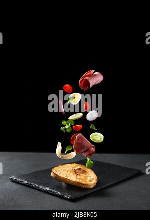 Ein Sandwich auf schwarzem Hintergrund. Die Zutaten fliegen in der Luft. Leerer Raum für Text. Sandwich mit Vetchina und Gemüse. Stockfoto