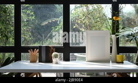 Bilderrahmen, Kaffeetasse, Bücher und eine Plättchenschale auf einem weißen Tisch in der Nähe eines Glasfensters im Home Office. Stockfoto