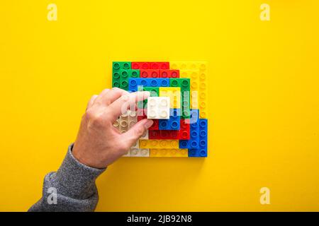 Hand, die einen weißen Spielzeugstein auf bunte Plastikbausteine legt und so eine Struktur erschafft Stockfoto