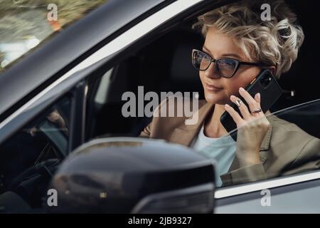 Elegante Frau in Brille, die während der Fahrt mit dem Auto telefonische Gespräche führt, Seitenansicht vom Fenster Stockfoto
