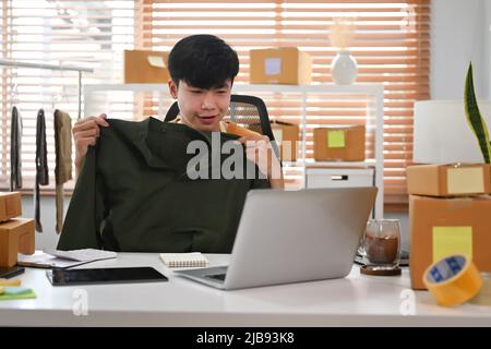 Junger asiatischer Mann zeigt Kleidung vor dem Laptop-Computer, während er Vlog-Videos und Live-Streaming online aufzeichnet. Stockfoto