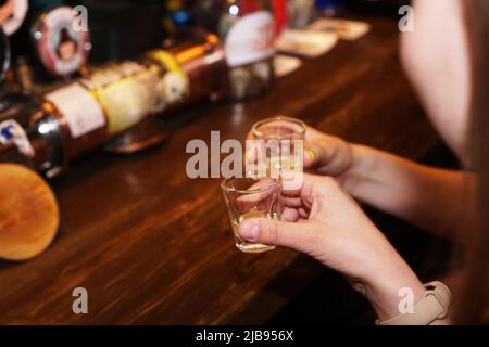 Zwei weibliche Hände klinken Gläser mit alkoholischen Schüssen. Unscharfer Balkenhintergrund. Gemütliche Bar vor Ort. Hintergrund für Menü und Alkoholkarte Stockfoto