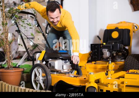 Fröhlicher Mann, der sich für den besten Rasenmäher im Gartengerätehaus entscheidet Stockfoto